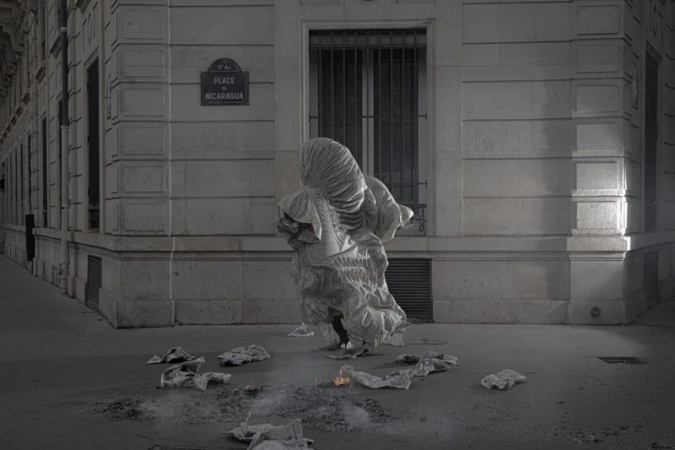 Model completely covered in a voluminous draped white gown standing on a street corner near scattered paper embers.