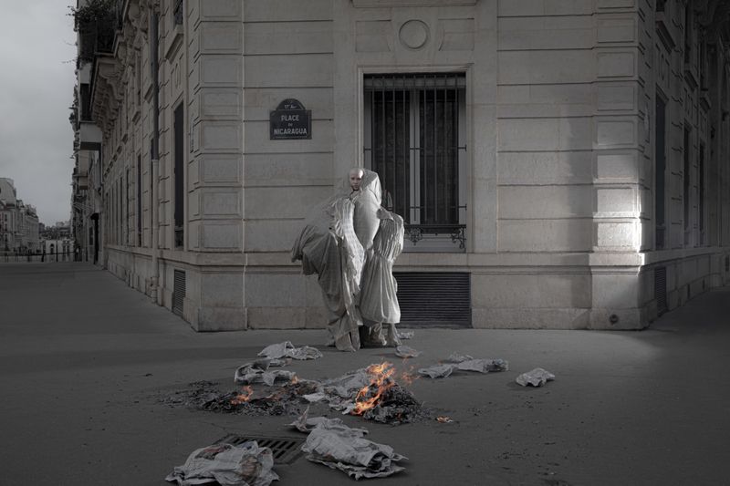 Model wrapped in voluminous draped fabric standing on a street corner next to burning newspapers outside a stone building.