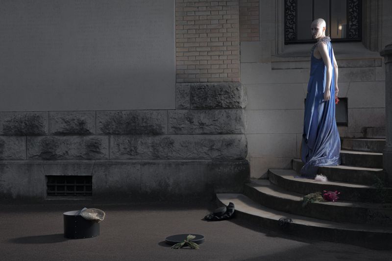 Model in long blue dress standing on stone steps outside a brick building with objects scattered on the ground.