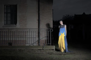 Model with a shaved head in a blue and yellow star-patterned gown standing beside a brick building at night.