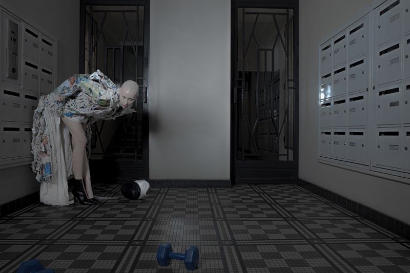 Model Victoria Cassie leaning forward in an avant-garde fringed coat in an apartment hallway with mailboxes and dumbbells.