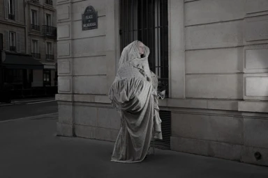 Model draped in heavy patterned fabric standing on a street corner against a classical stone building facade in a monochrome photograph.