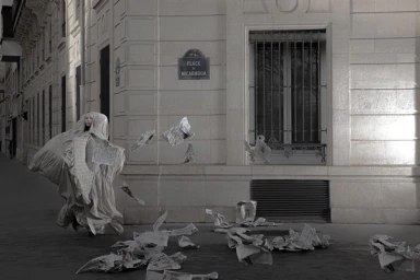 Model Victoria Cassie in an oversized hooded draped coat surrounded by flying newspapers on a Paris street.