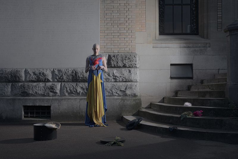 Model with a shaved head standing outdoors in a draped blue, yellow, and red gown beside stone steps.