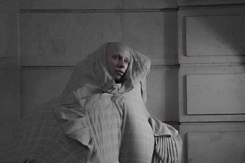 Bald model Victoria Cassie wrapped in voluminous draped plaid fabric against an architectural stone wall.