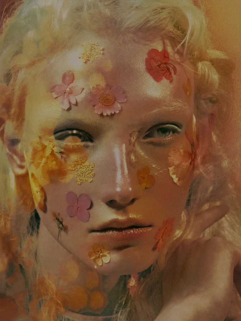 Close-up portrait of a blonde model with colorful pressed flowers applied across her face in soft, warm lighting.