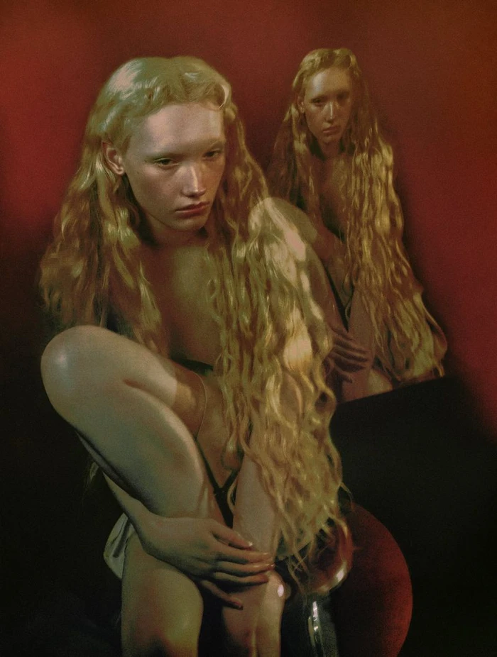 Model Ivy Stewart with long crimped blonde hair crouches in a warm, painterly red studio setting photographed by Elizaveta Porodina.