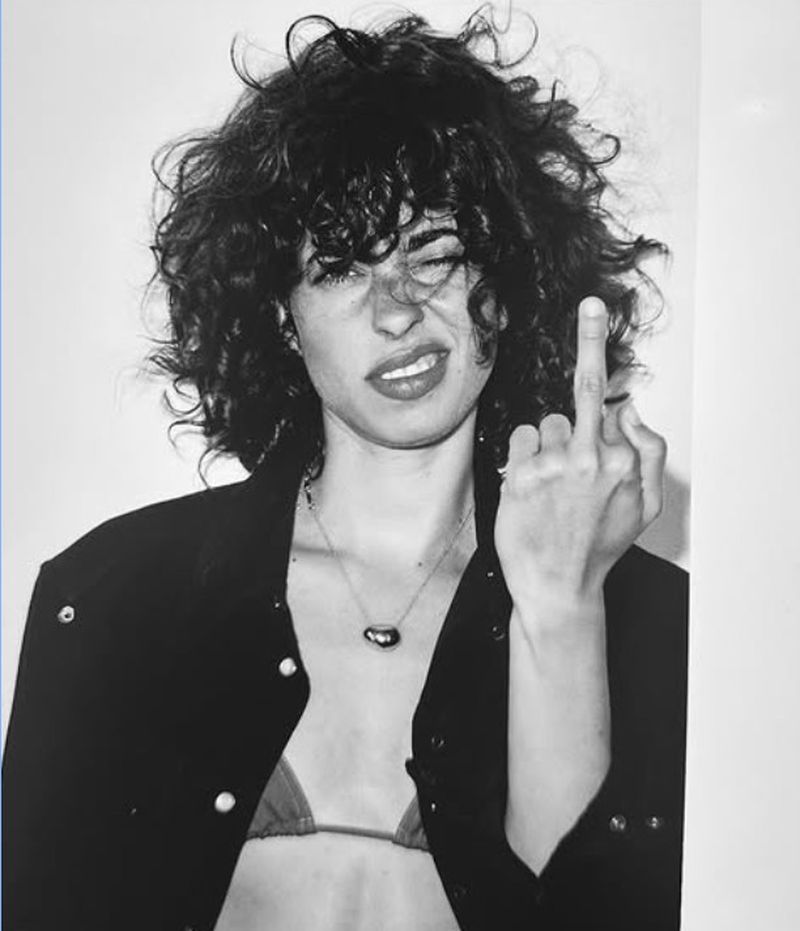 Black and white portrait of a young woman with curly hair wearing a jacket and necklace, extending her middle finger toward the viewer.
