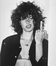 Model Marina Ontanaya with curly dark hair winks and flips the middle finger in a black jacket and necklace against a white background.