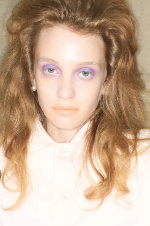 Close-up beauty portrait of model Dasha Page with voluminous wavy hair, purple eyeshadow, and pale pink collared shirt.