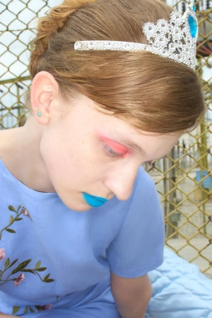 Model wearing a blue t-shirt and sparkly tiara with a blue gemstone outdoors against a wire fence