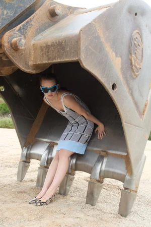 Model wearing patterned dress and blue sunglasses posed inside a large excavator bucket on sandy ground