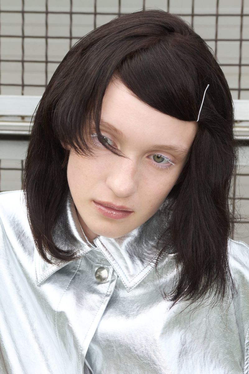 Model Dasha Page wearing a metallic silver jacket with styled dark hair secured by a pin against a wire grid background.