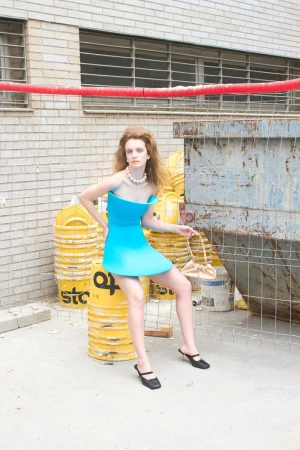 Model Dasha Page in a bright cyan structured mini dress seated on yellow buckets next to an industrial dumpster.