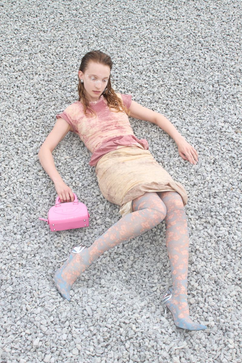 Model reclined on light grey gravel wearing a layered pink dress and holding a small bright pink handbag.