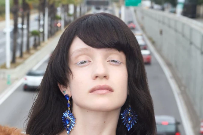Model Dasha Page with dark fringe bangs, white eyeliner, and blue crystal chandelier earrings against a blurred highway background.
