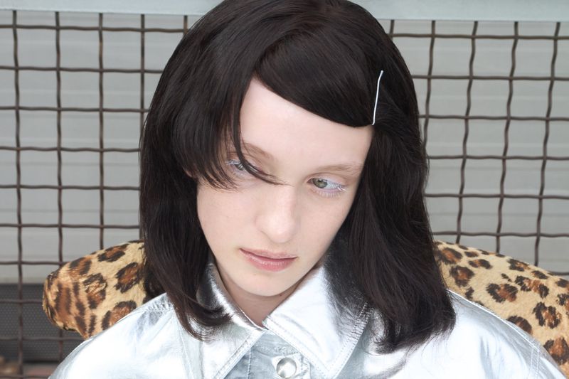 Dasha Page wearing a silver metallic collared shirt seated against a leopard print chair and wire mesh grid.