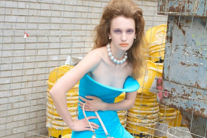 Model with bouffant hair in a sculptural blue strapless dress and oversized pearl choker posed behind a wire fence.