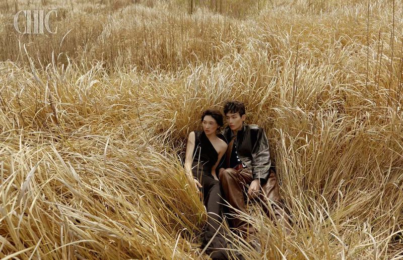 A woman in a black dress and a man in a leather jacket recline together amidst tall, dry golden grass.