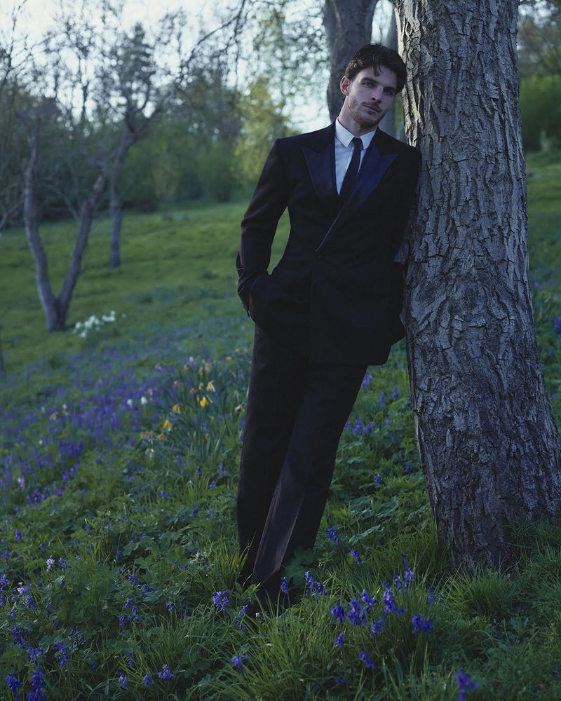 Declan Rice wearing a black double-breasted suit leaning against a tree trunk in a field of wildflowers.