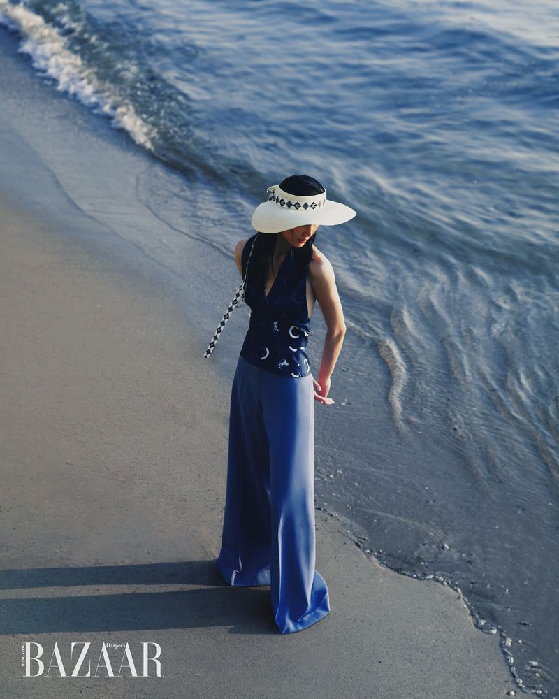 Du Si Ying wears blue wide-leg trousers, a navy patterned halter top, and a white sun visor on a sandy beach by the ocean water.
