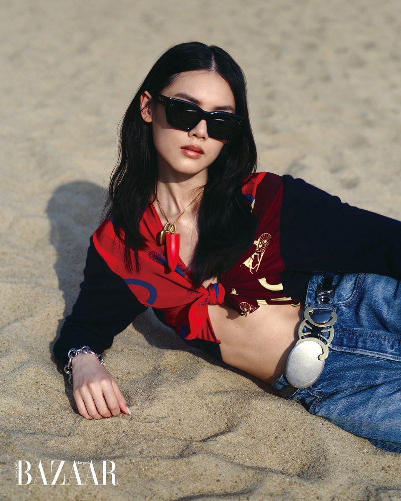 Model Du Si Ying wearing dark sunglasses, a red patterned cropped shirt, and blue denim jeans reclining on sand.