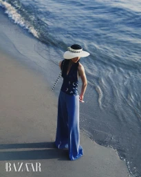 Du Si Ying wears blue wide-leg trousers, a navy patterned halter top, and a white sun visor on a sandy beach by the ocean water.