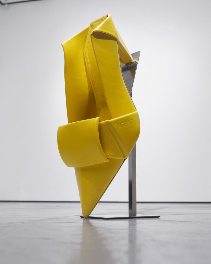 Sculptural yellow leather pointed-toe shoe mounted vertically on a metal display stand in a white gallery space.
