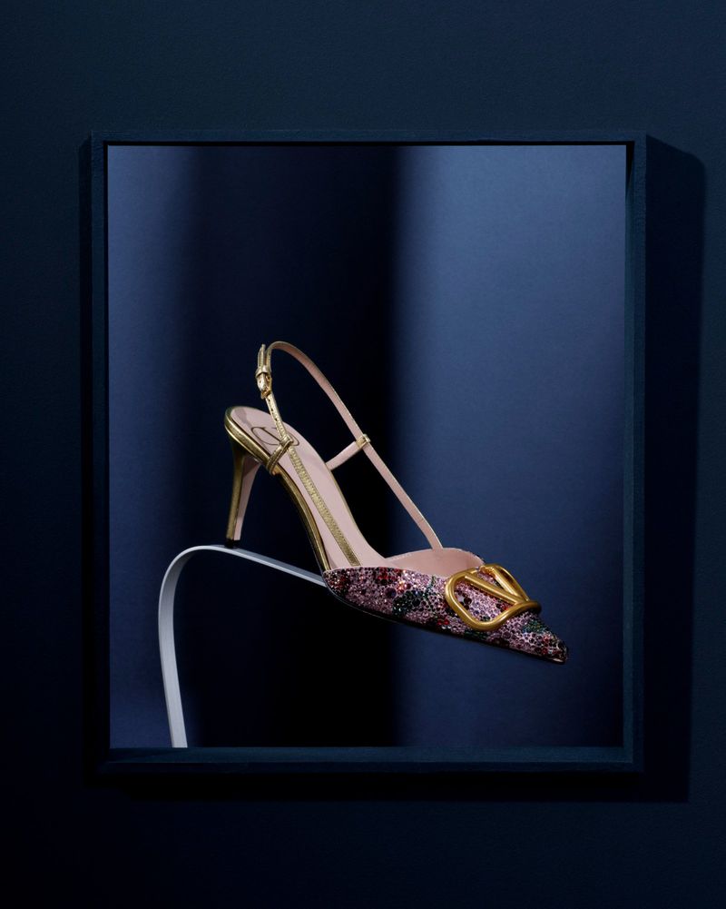 Embellished pointed-toe slingback heel with gold buckle displayed on a curved stand inside a navy frame.
