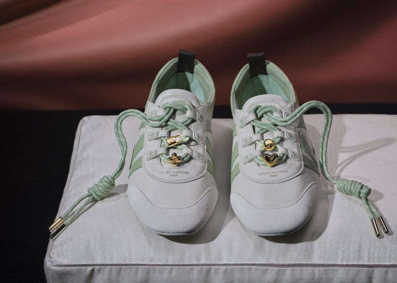 Pair of white and pale green designer sneakers with gold charms on light green cord laces resting on a white cushion.