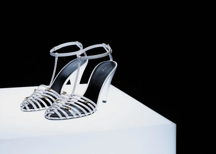 Pair of white strappy high-heeled sandals with black insoles displayed on an illuminated white pedestal against a solid black background.