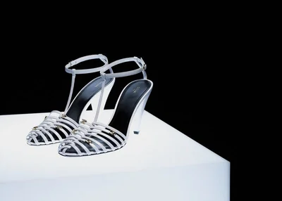 Pair of white strappy high-heeled sandals with black insoles displayed on an illuminated white pedestal against a solid black background.