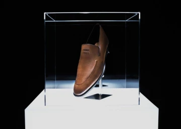 A brown leather loafer exhibited inside a clear glass vitrine on an illuminated white pedestal against a dark backdrop.