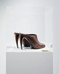 Brown leather heeled mules displayed inside a clear acrylic museum vitrine on a white pedestal against a pale studio backdrop.