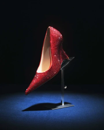 A red crystal-embellished pointed-toe pump mounted on a metal display stand under a direct spotlight on a dark blue surface.