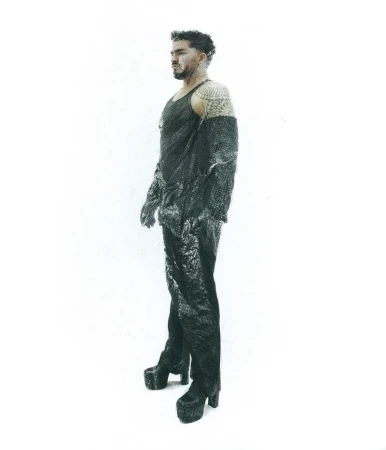 Adam Lambert posing in a dark textured sleeveless outfit and platform boots against a white studio background.