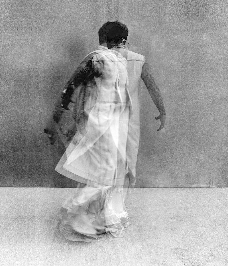 Black and white multiple exposure photograph of Adam Lambert standing against a textured wall wearing a draped sleeveless garment.