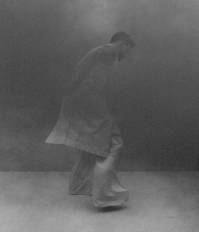 Adam Lambert in a long sleeveless coat and wide-leg trousers walking through a hazy monochrome studio.