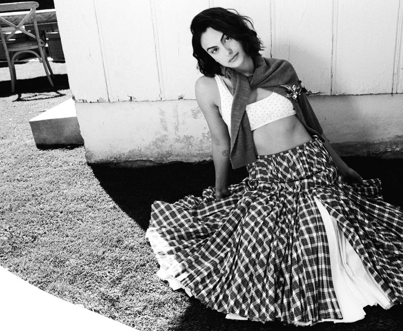 Camila Mendes seated outdoors wearing a plaid skirt, white crop top, and a sweater draped over her shoulders.