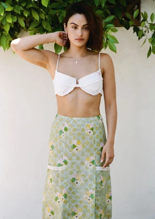 Camila Mendes poses in a white bralette top and patterned green midi skirt against a pale wall with leafy foliage above.