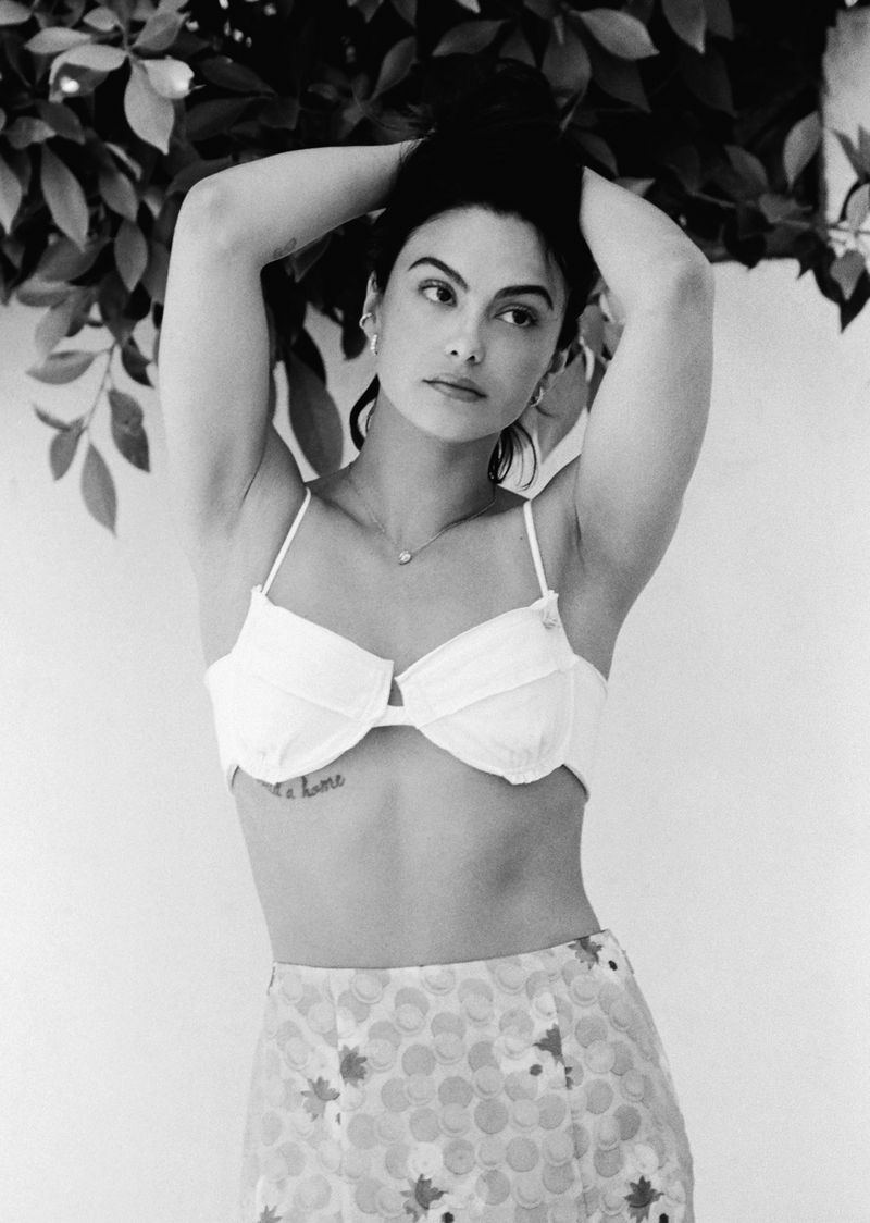 Black and white portrait of Camila Mendes posing with hands behind her head wearing a white bralette and patterned skirt beneath foliage.