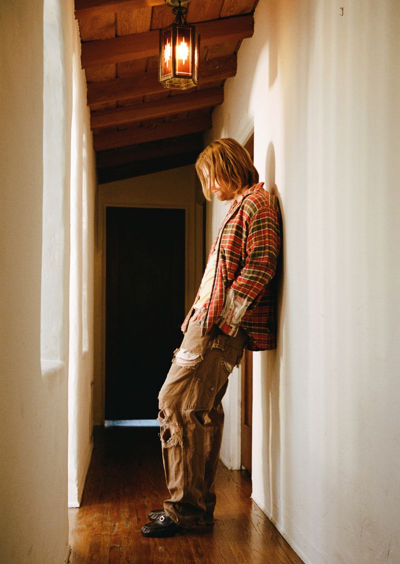 Finneas leans against a white hallway wall wearing a red plaid shirt and distressed brown cargo pants.