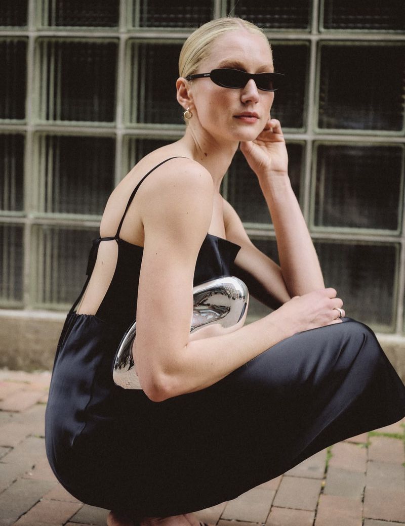 A woman crouching outdoors in a black backless slip dress and dark narrow sunglasses, holding a sculptural silver clutch.