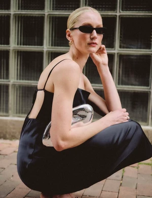 A woman crouching outdoors in a black backless slip dress and dark narrow sunglasses, holding a sculptural silver clutch.