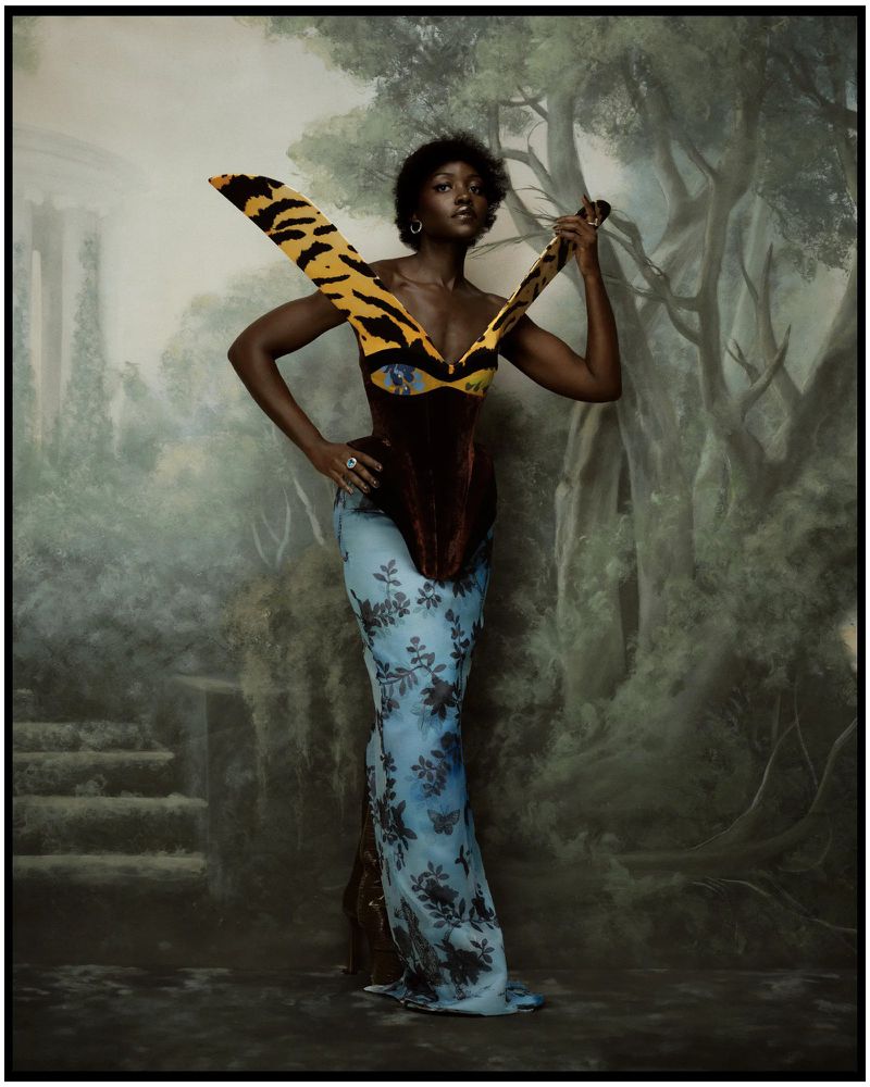 Lupita Nyong'o poses in a structured gown with winged tiger-print collar and floral blue skirt before a painted woodland backdrop.