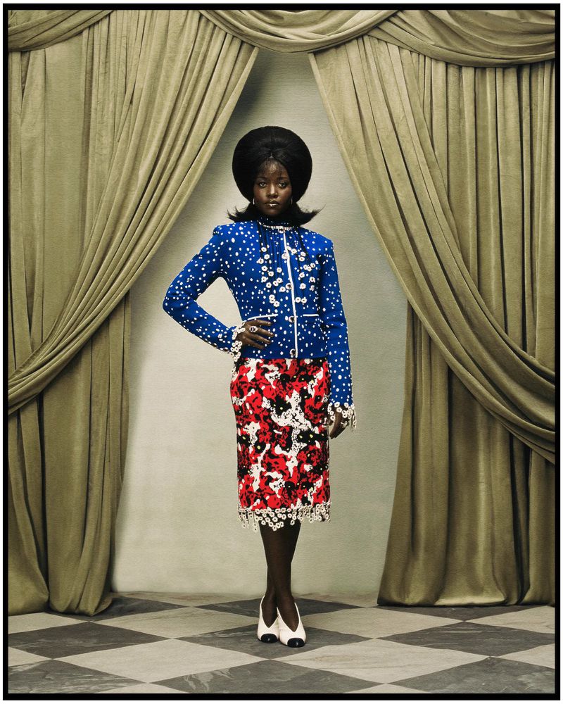 Lupita Nyong'o in a blue patterned jacket and red skirt standing framed by heavy stage curtains.