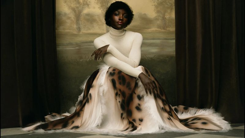 Lupita Nyong'o seated in a white turtleneck and sweeping spotted skirt in front of a painted landscape backdrop.