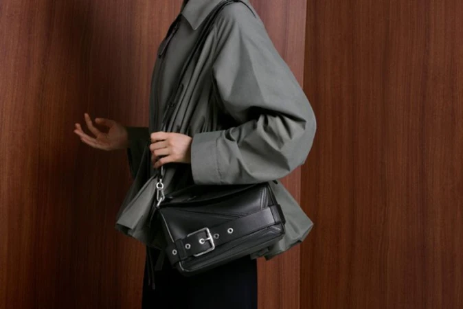 Person wearing an oversized greyish collared jacket carrying a black leather crossbody bag with a large silver buckle against wood panels.