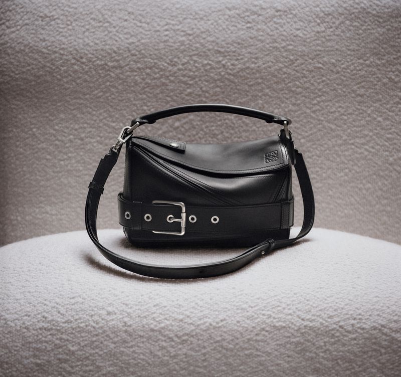 Elegant black leather Loewe bag with a buckle strap, presented on a soft, textured light-grey surface in a studio setting.