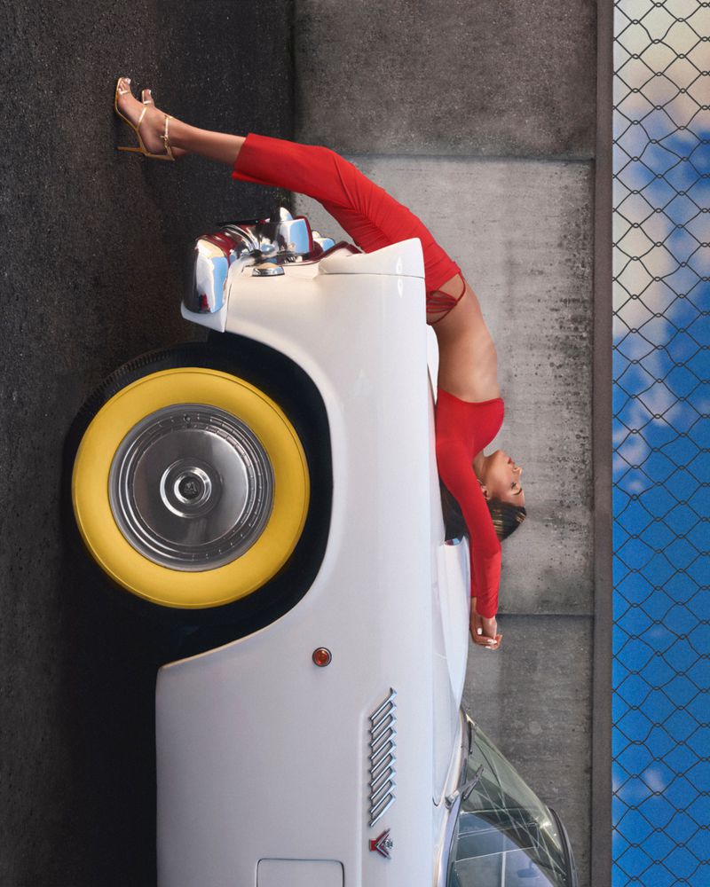 An adult woman in red pants and a top leans upside down on a white classic car. A chain link fence and blue sky in the background.